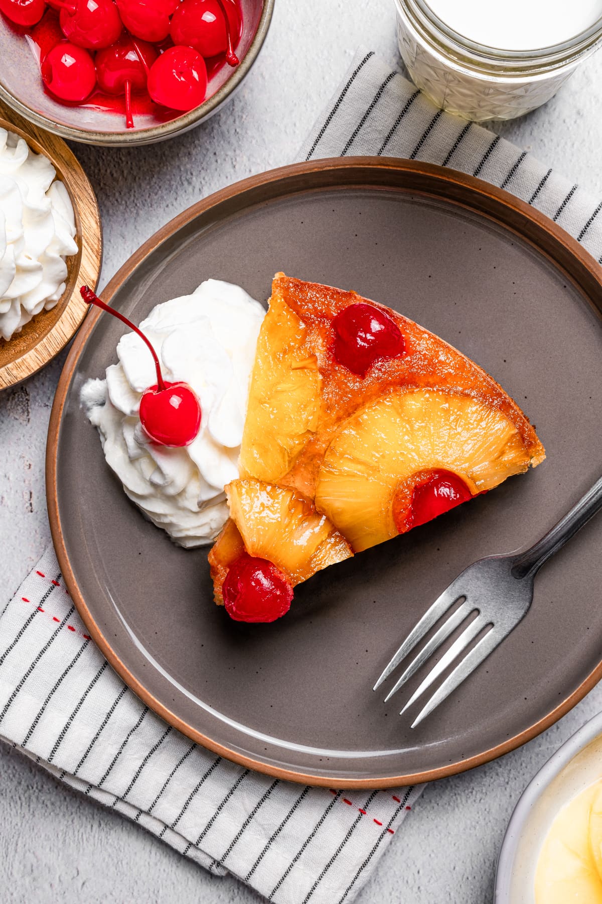 Slice of pineapple upside down cake on a stoneware plate, next to a fork and a swirl of whipped cream topped with a cherry.