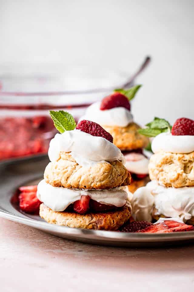 Three strawberry shortcakes desserts served on a pewter plate with a bowl of strawberries behind them