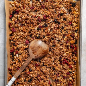 Overhead image of homemade granola on a baking sheet with a wooden spoon stirring through it.