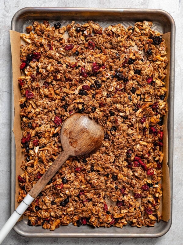 Overhead image of homemade granola on a baking sheet with a wooden spoon stirring through it.