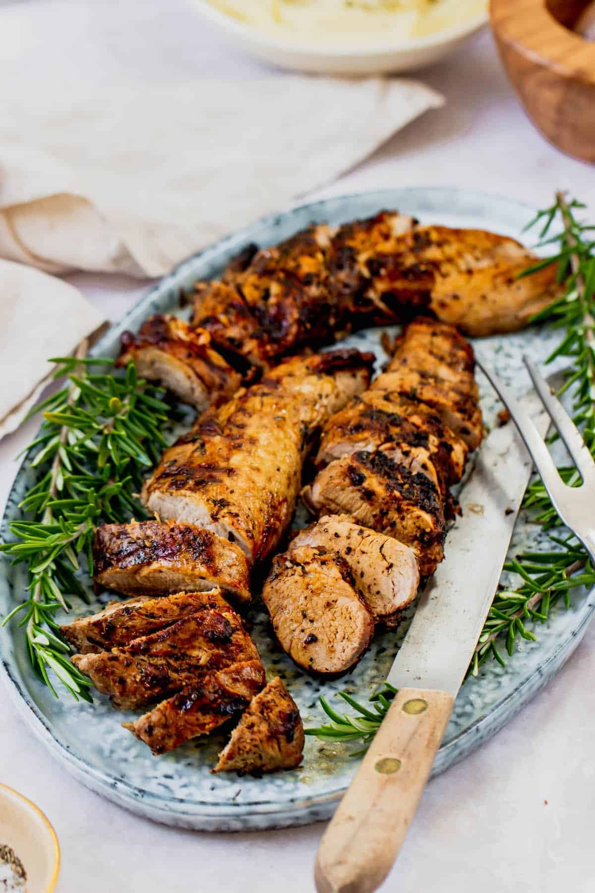 A platter with two grilled pork tenderloins, fresh rosemary, and serving utensils.