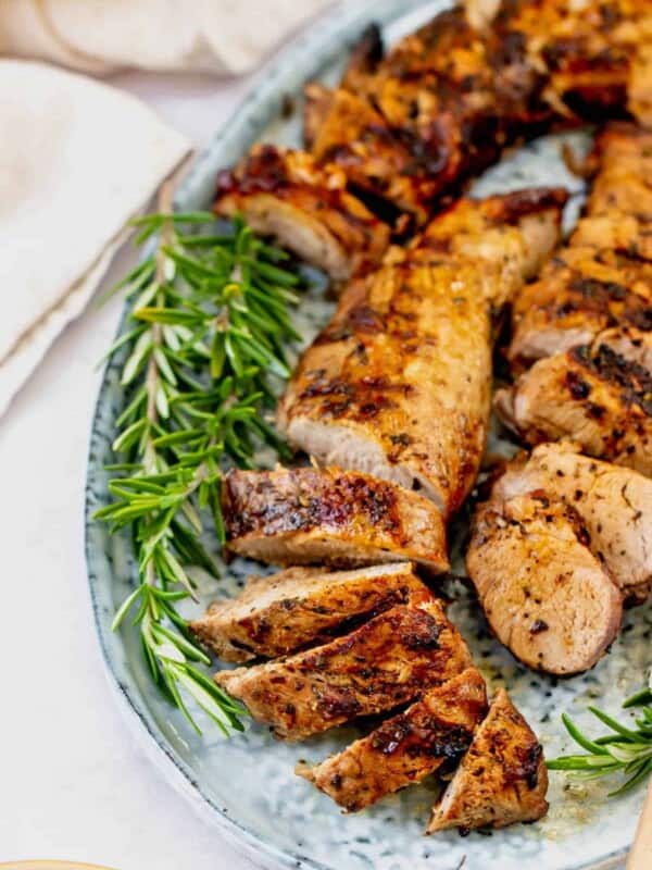 Close-up of grilled pork tenderloin on a serving platter, garnished with fresh rosemary. Some of the pork is sliced into medallions.