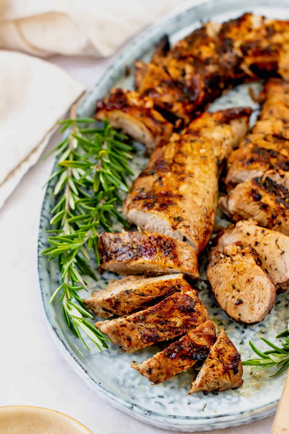 Close-up of grilled pork tenderloin on a serving platter, garnished with fresh rosemary. Some of the pork is sliced into medallions.