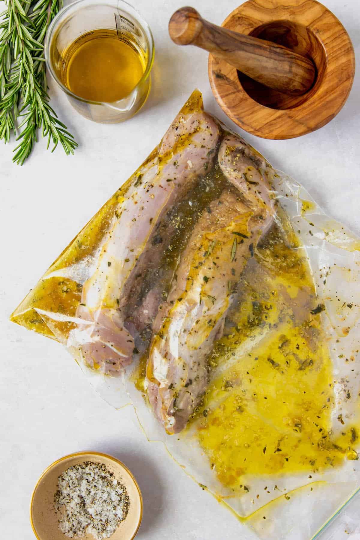 Uncooked pork marinating in a ziploc bag. Olive oil, herbs, a mortar and pestle, and salt are on the work surface nearby.