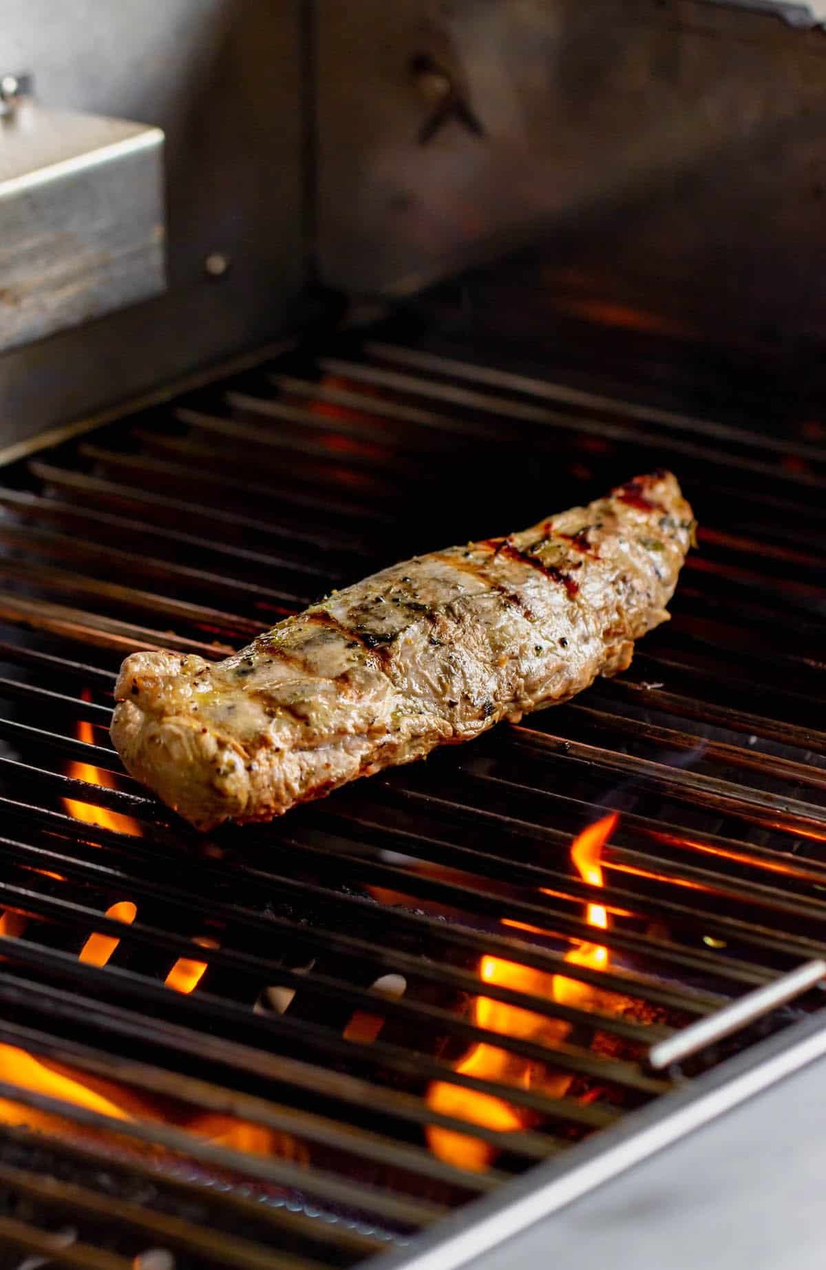 A whole pork tenderloin on a grill. Flames are visible under the grill grates.