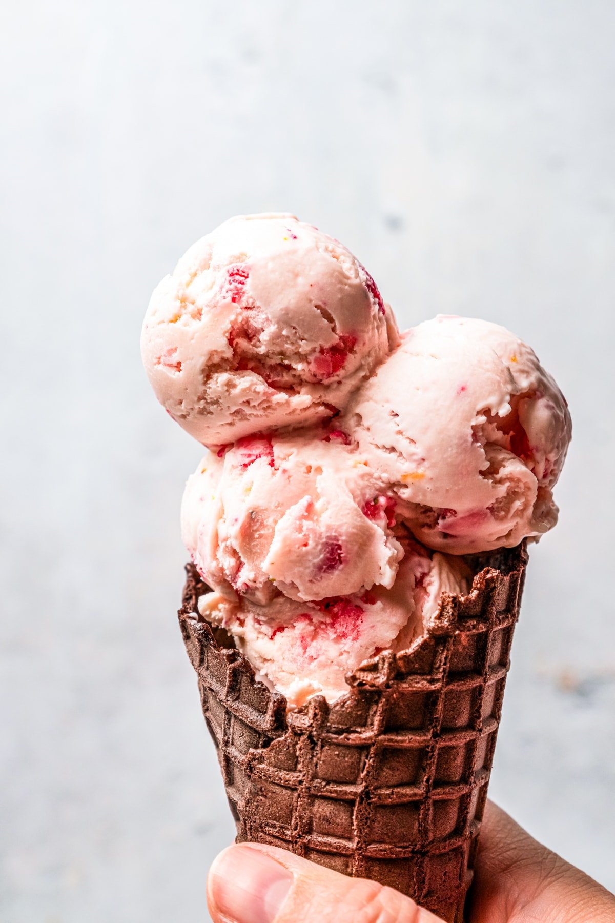 Hand holding up a waffle cone filled with three scoops of strawberry cheesecake ice cream.