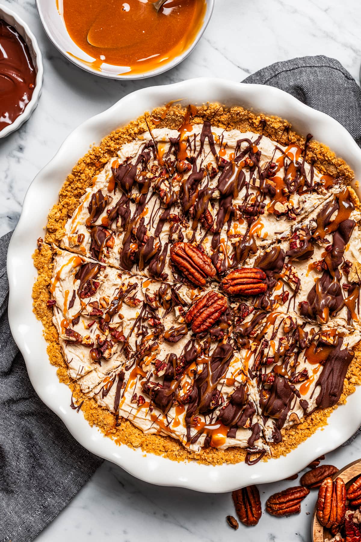 Overhead view of Turtle pie topped with toasted pecans and drizzled with caramel and chocolate, next to bowls of toppings.