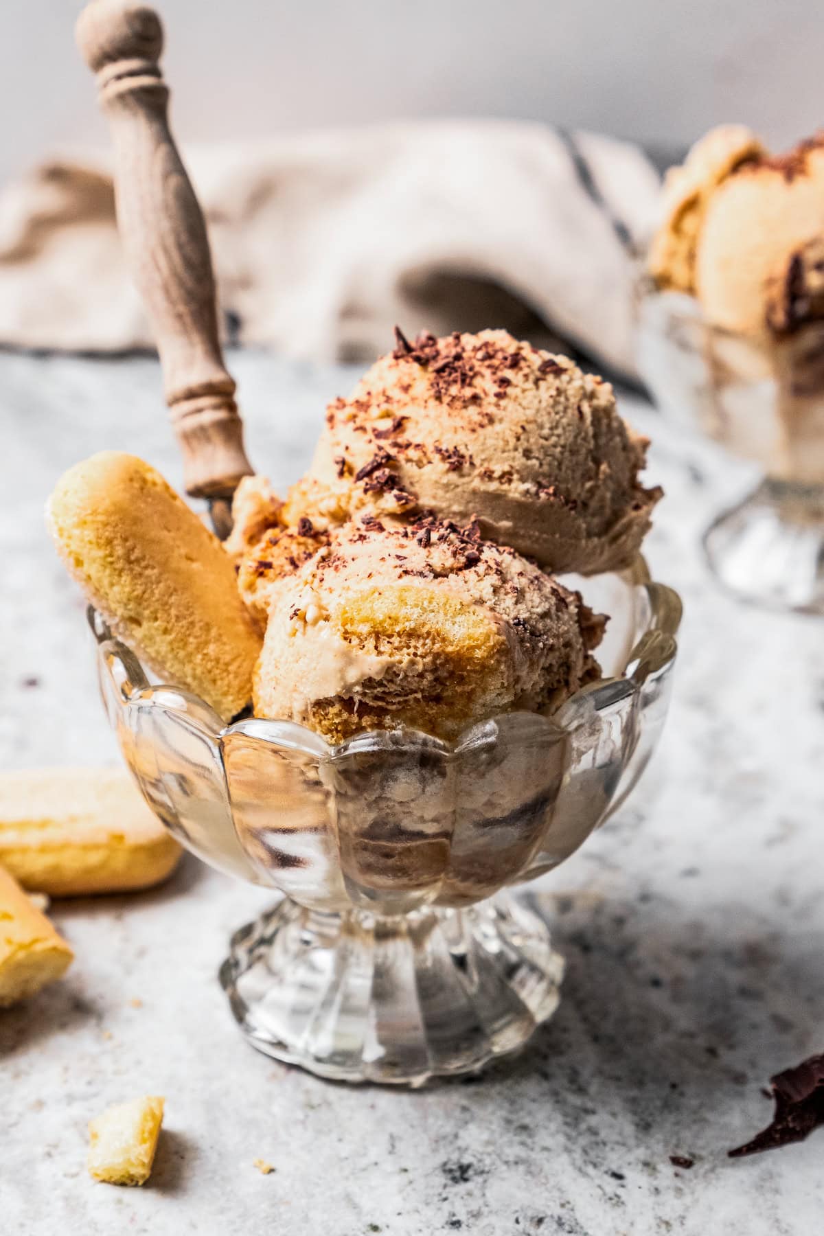 Scoops of tiramisu ice cream in glass bowls, next to scattered ladyfinger cookies and chopped chocolate.