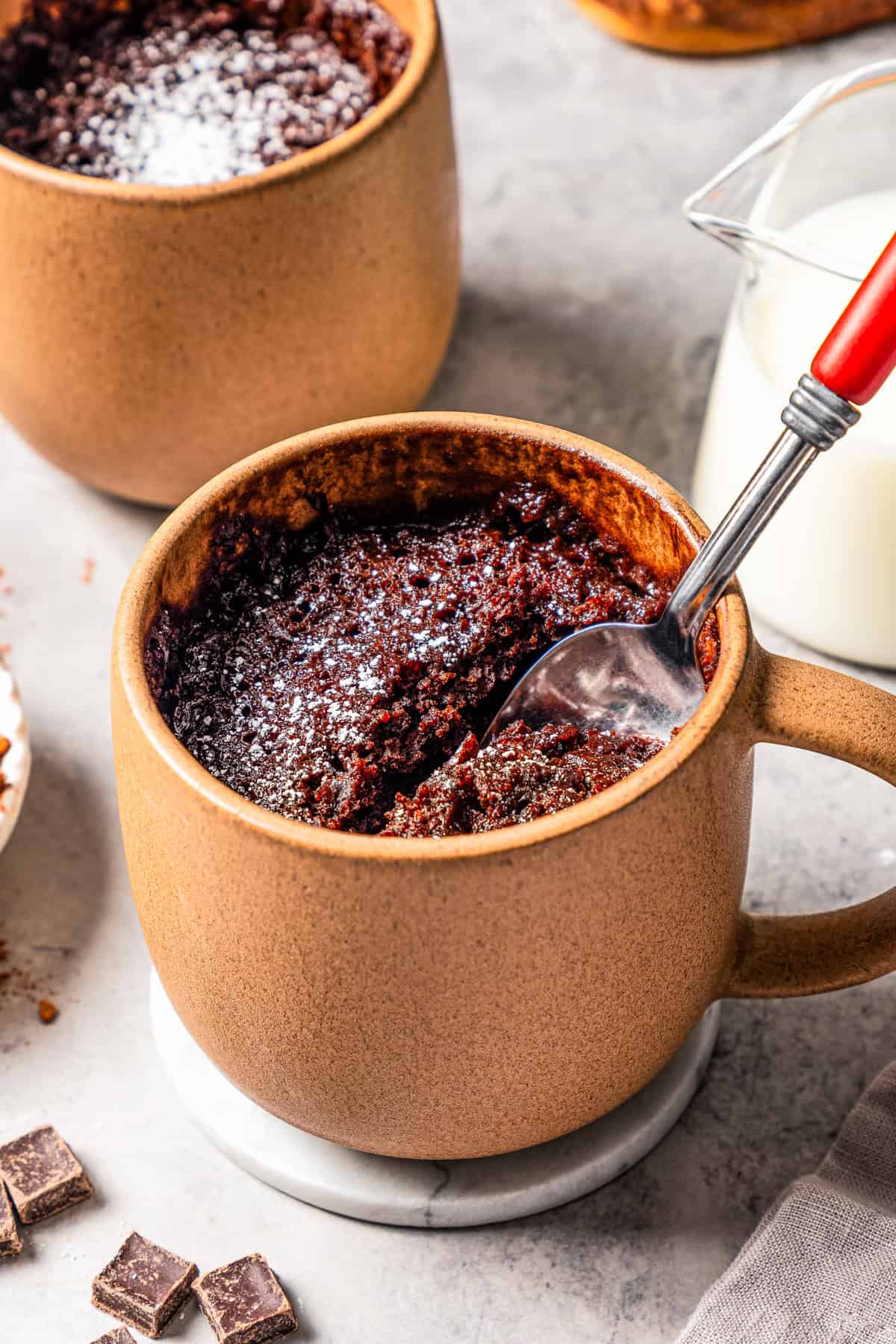 A spoon in a chocolate mug cake, with a second mug cake in the background.