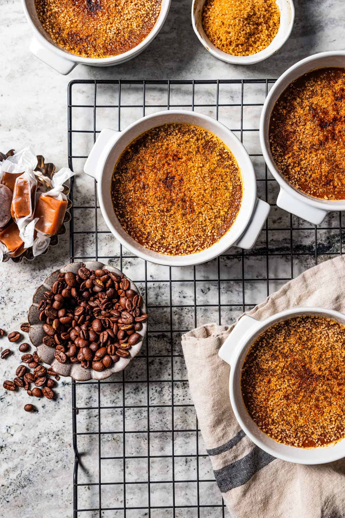 Crème brûlée in ramekins next to small bowls of coffee beans and caramels on a wire rack.