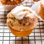 An apple pie muffin topped with cinnamon sugar and vanilla glaze on a wire rack, with more muffins in the background.