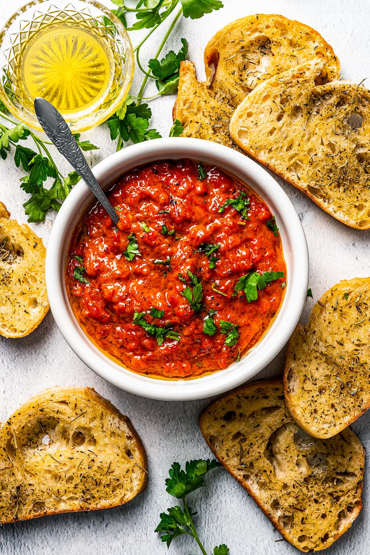 Ajvar in a white dish surrounded by bread slices and a small bowl of olive oil.