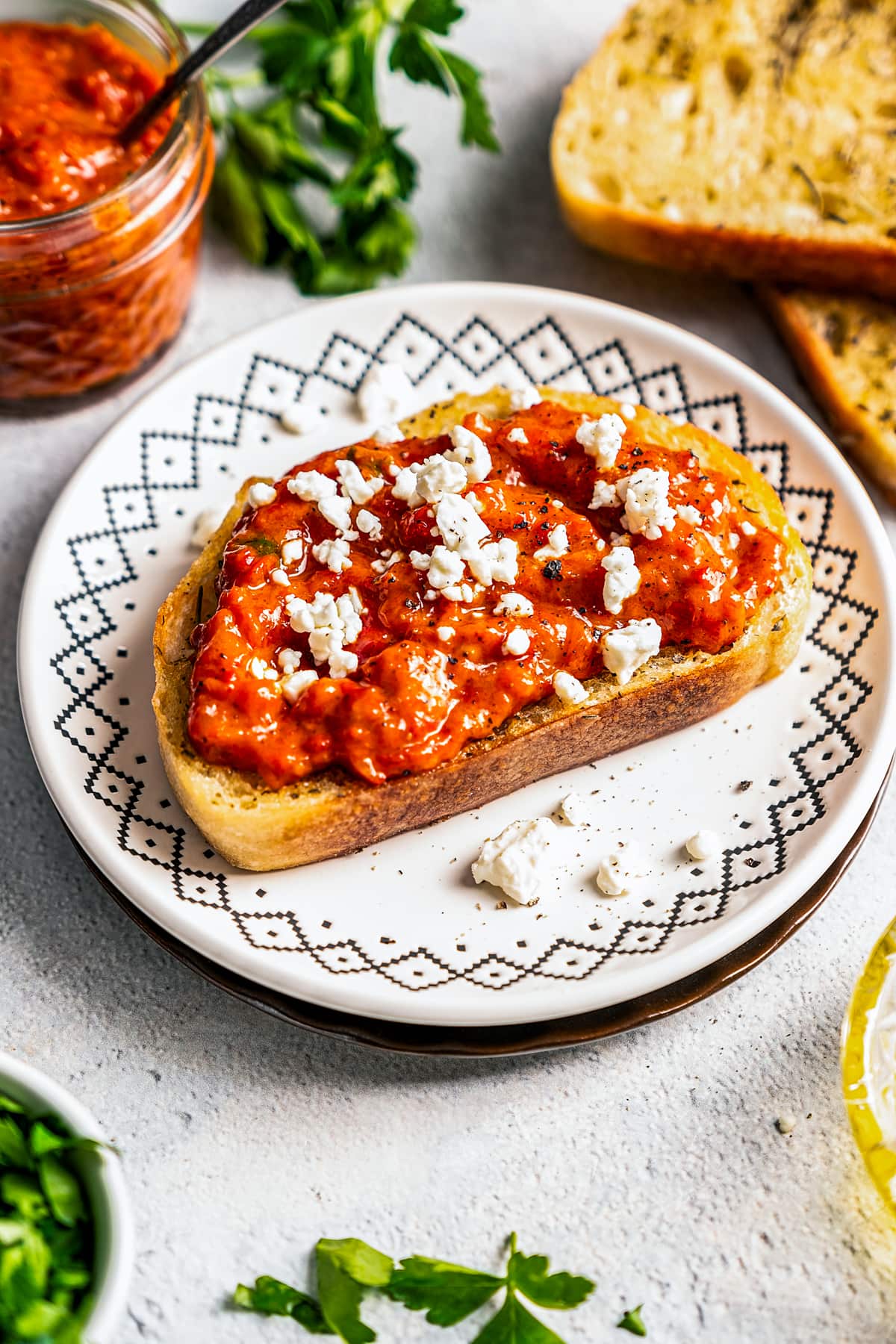 Red pepper relish spread over a piece of toasted bread, topped with crumbled feta, served on a plate, and set next to a jar of Ajvar and more toast slices.