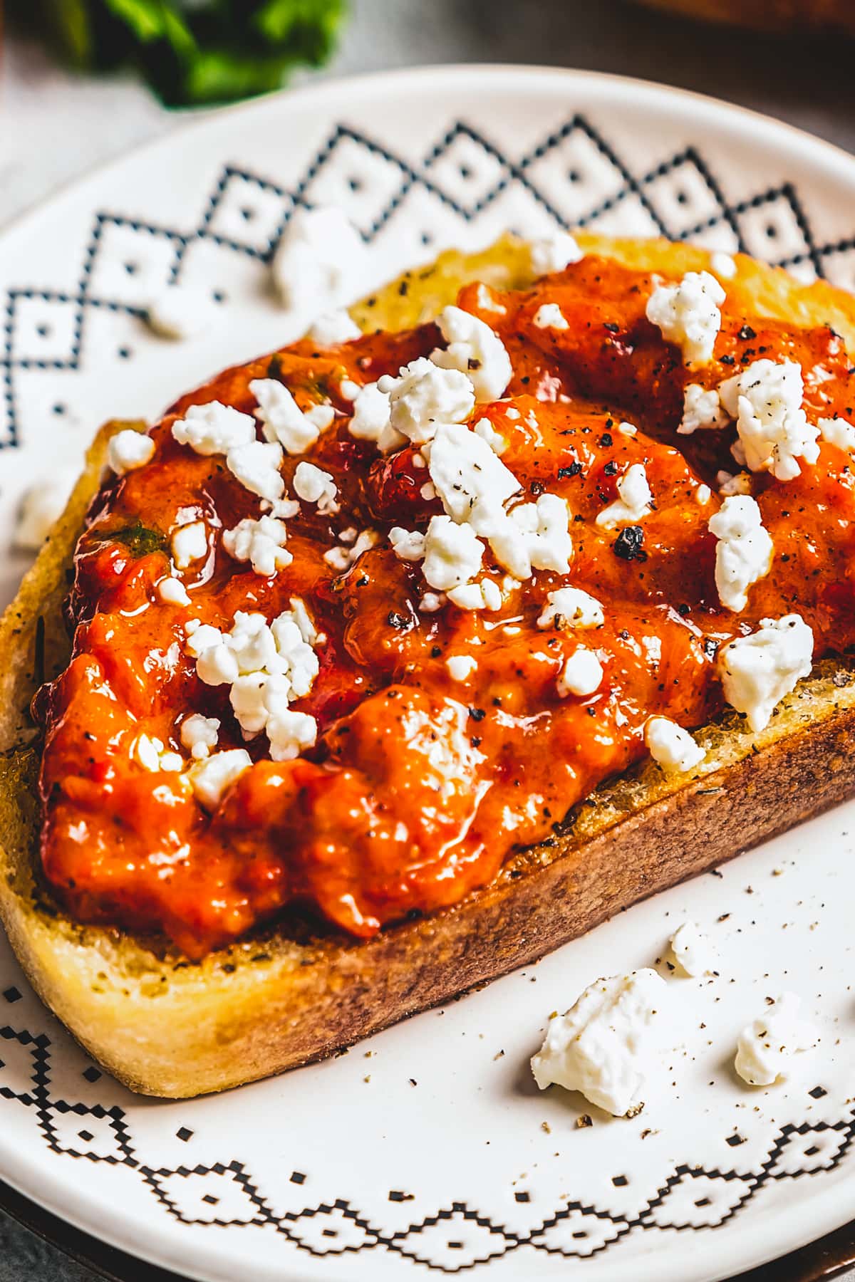 Red pepper relish spread over a piece of toasted bread, topped with crumbled feta, and served on a plate.