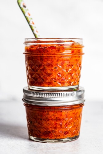 Two small jars of Ajvar stacked, with a spoon sticking out of the top jar.