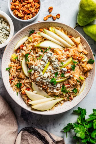 Overhead view of orzo salad with pears, walnuts and gorgonzola cheese in a bowl garnished with pear slices, with a wooden spoon, surrounded by bowls of ingredients.