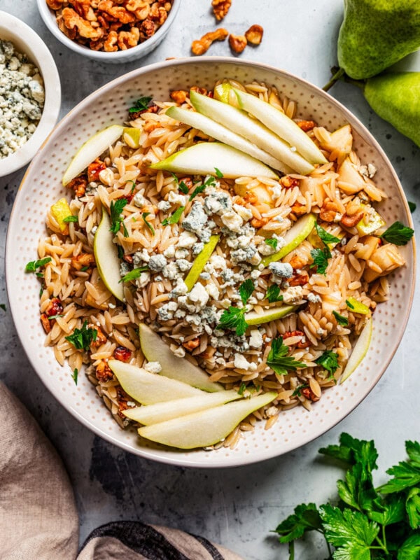 Overhead view of orzo salad with pears, walnuts and gorgonzola cheese in a bowl garnished with pear slices, with a wooden spoon, surrounded by bowls of ingredients.