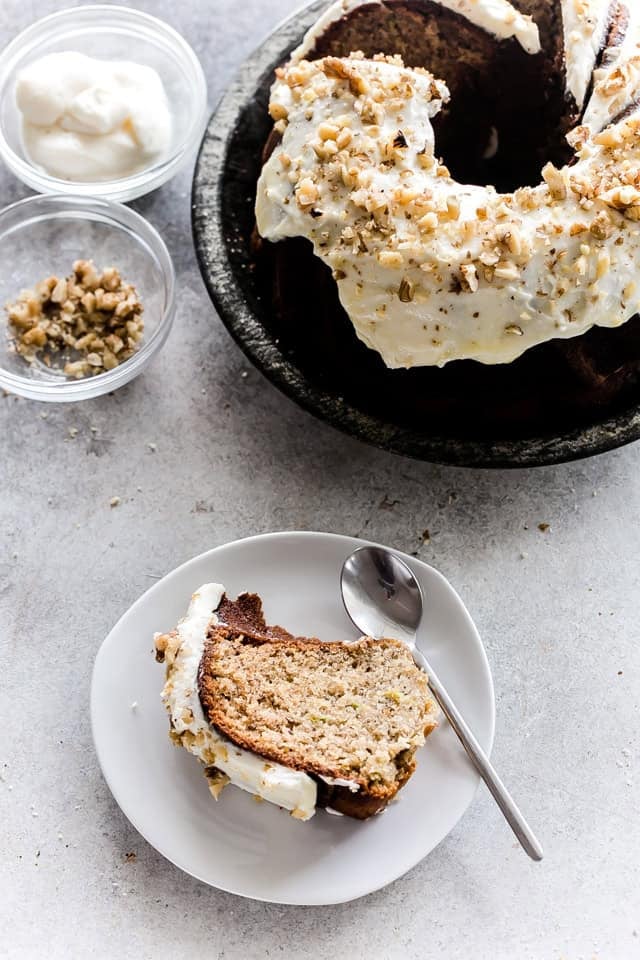 This moist banana zucchini cake is made with ripe bananas, fresh zucchini, and warm spices, then topped with maple cream cheese frosting and walnuts. Slice of banana zucchini cake with maple cream cheese frosting and walnuts beside the frosted Bundt cake.