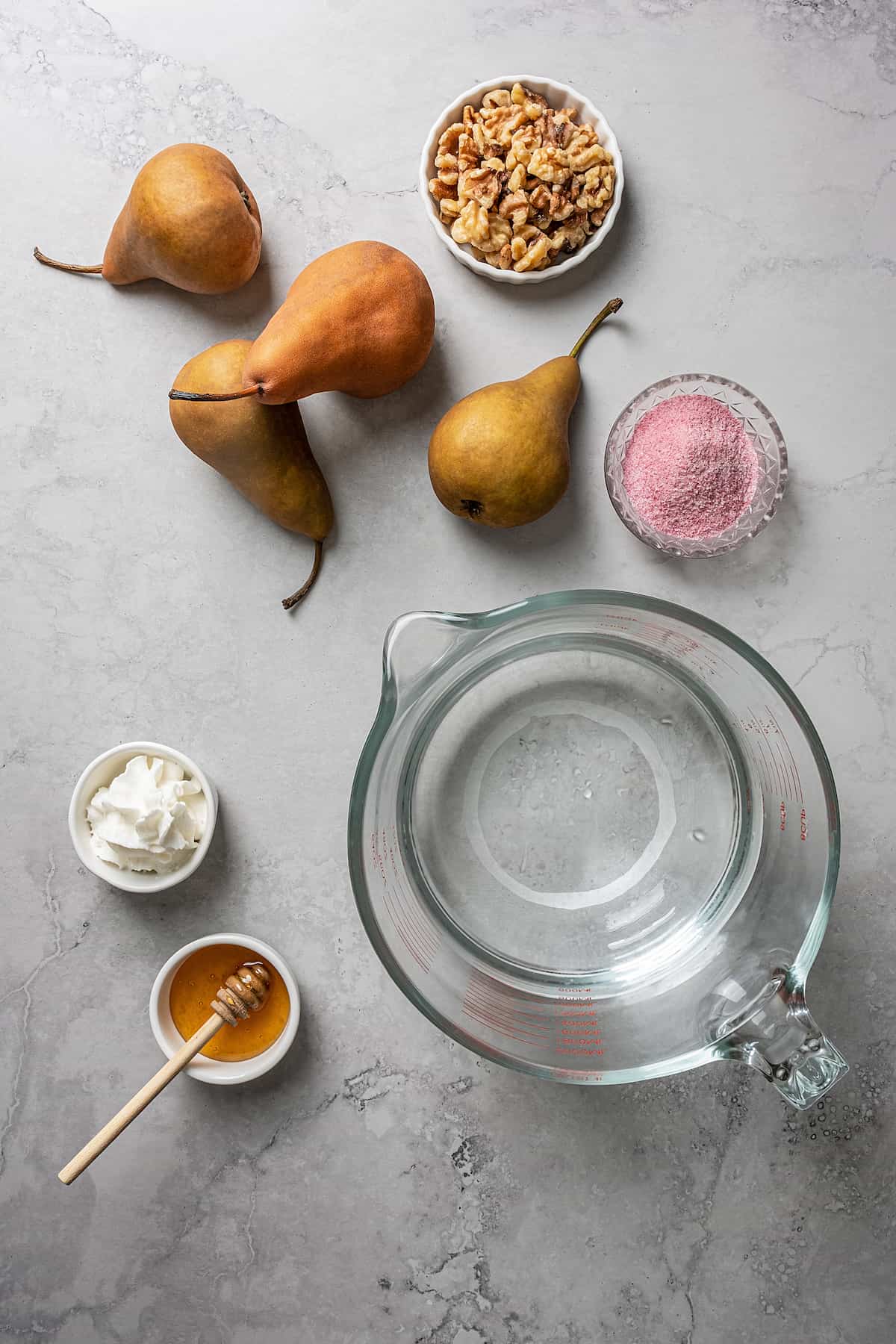 Overhead view of the ingredients needed for poached pears: four whole pears, a bowl of walnuts, a bowl of Jell-O gelatin, a bowl of whipped cream, a bowl of honey with a wooden honey spoon, and a pyrex of water