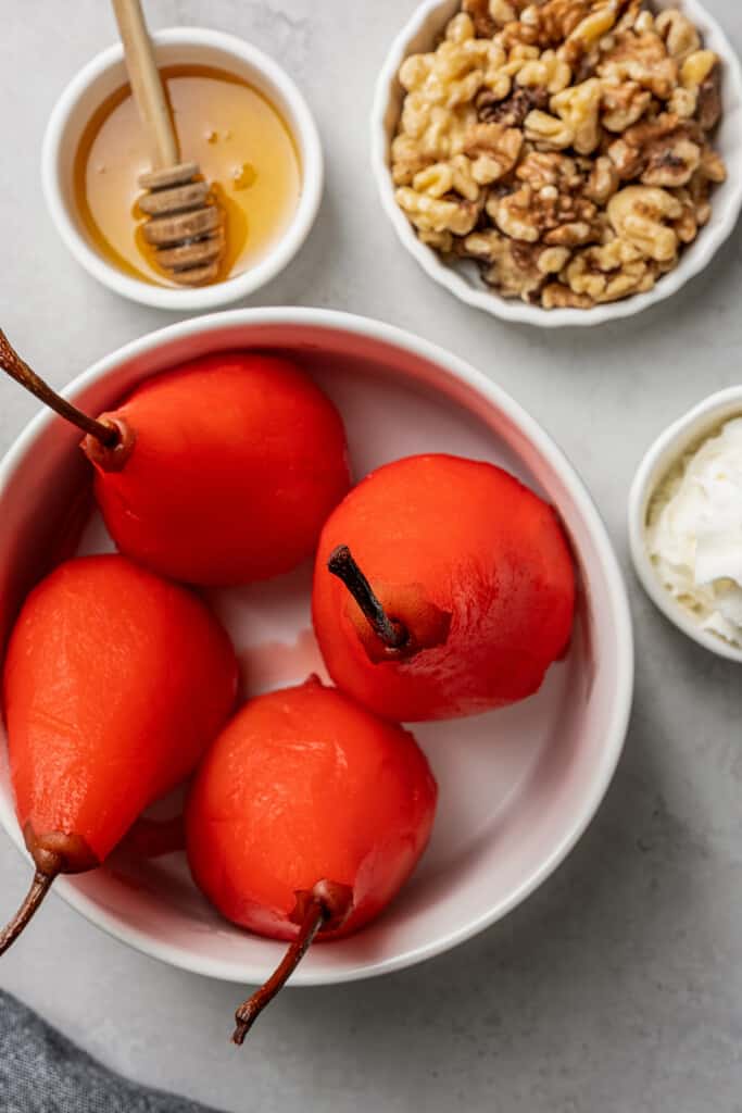 Close up overhead view of four poached pears in a bowl, next to a bowl of honey with a wooden honey spoon, a bowl of walnuts, and a bowl of whipped cream