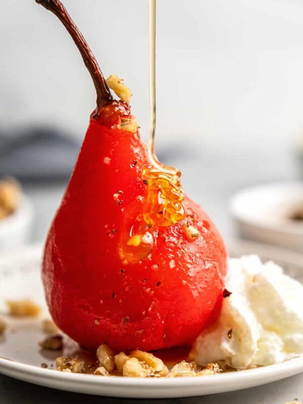 Close up of a poached pear on a plate with walnuts and whipped cream, with h oney being poured over it