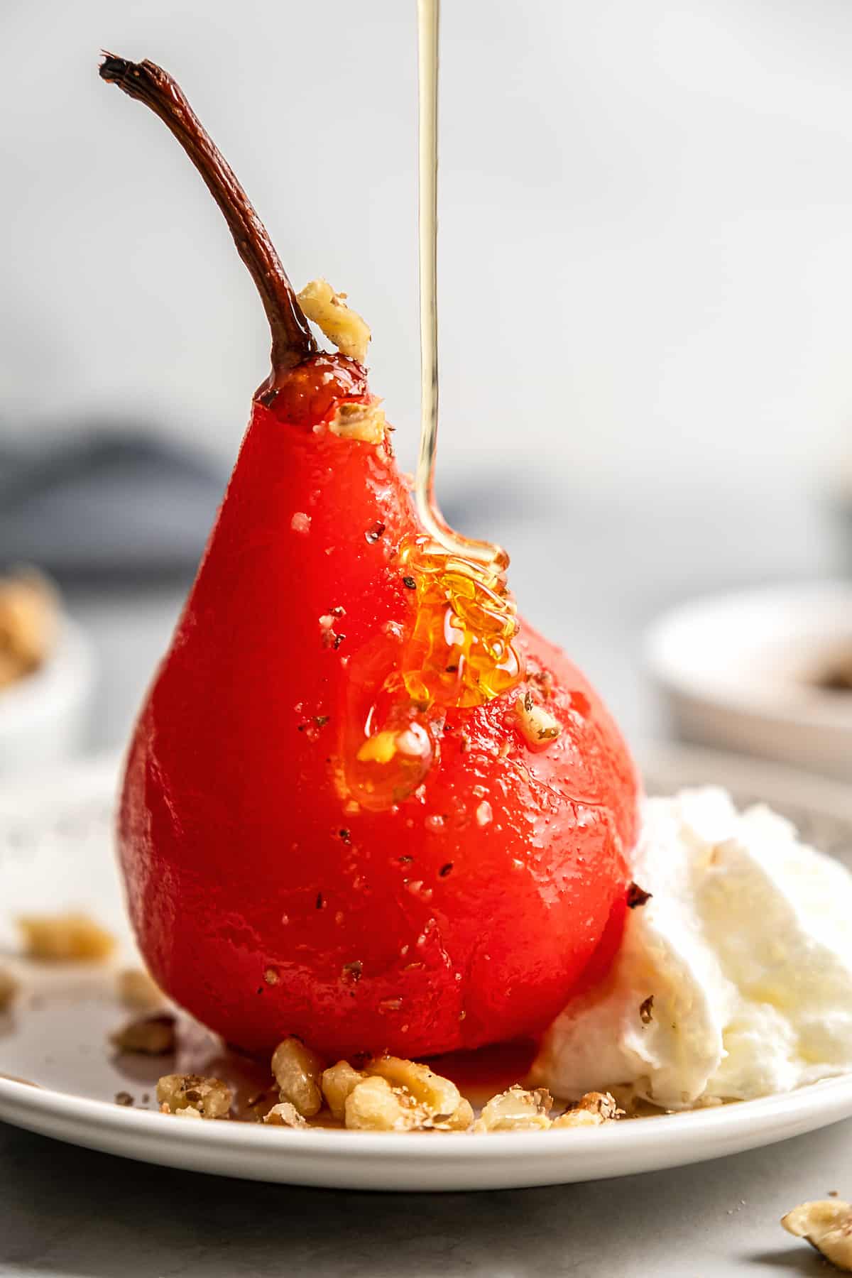 Close up of a poached pear on a plate with walnuts and whipped cream, with honey being poured over it