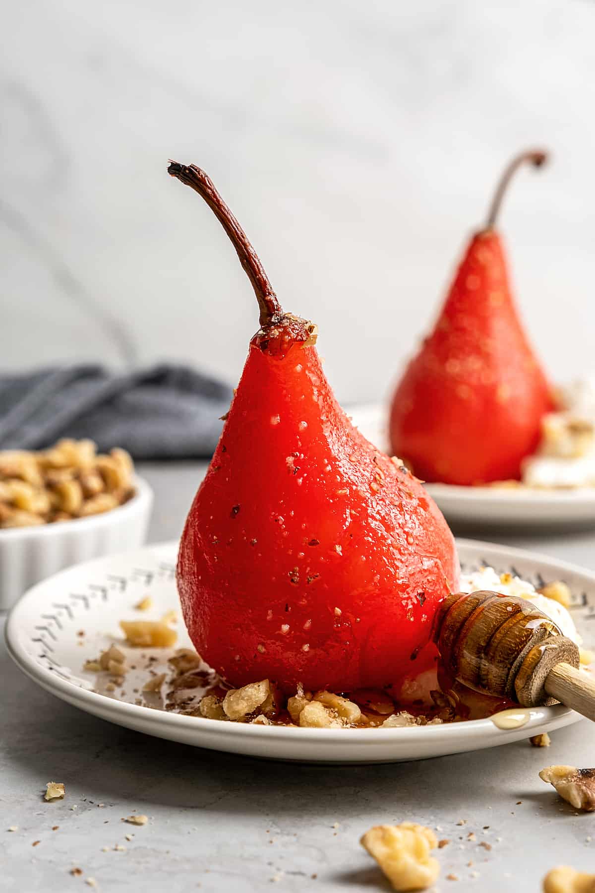 A poached pear on a plate with walnuts and a wooden honey spoon, with a bowl of walnuts and another poached pear in the background