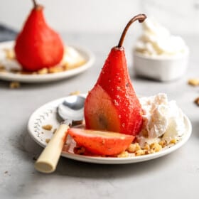 A poached pear that has been cut into on a plate with a spoon, walnuts, and whipped cream, with another poached pear and a bowl of whipped cream in the background