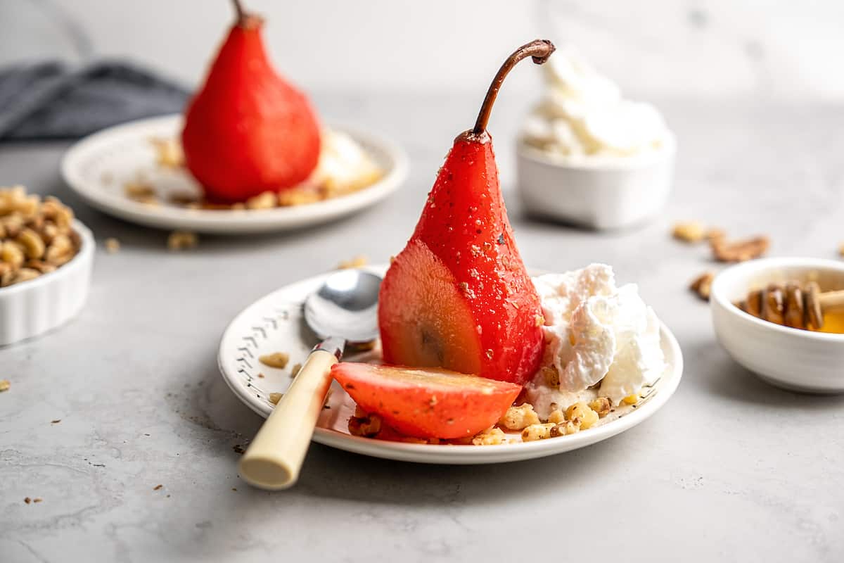 A poached pear that has been cut into on a plate with a spoon, walnuts, and whipped cream, with another poached pear and a bowl of whipped cream in the background