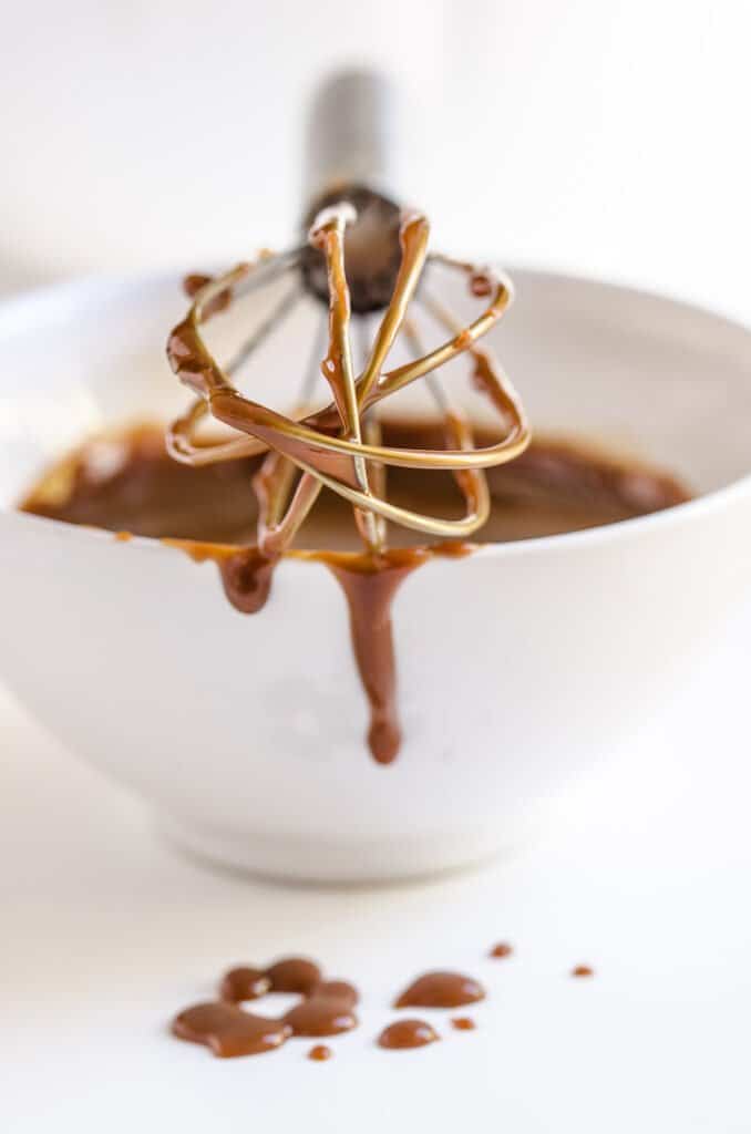 A whisk with melted caramel balanced on a white bowl