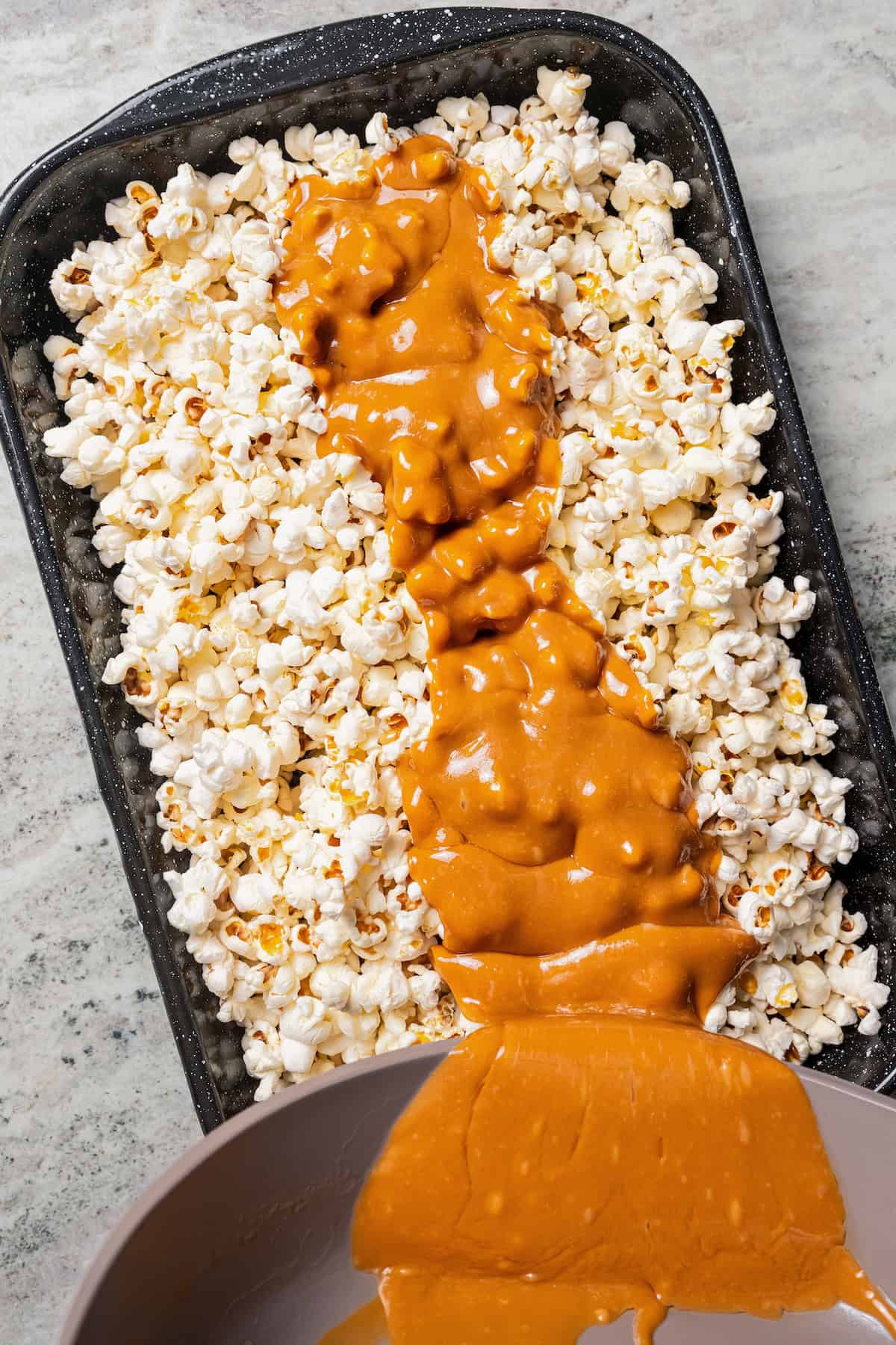 Pouring melted caramel over popcorn on a baking sheet.