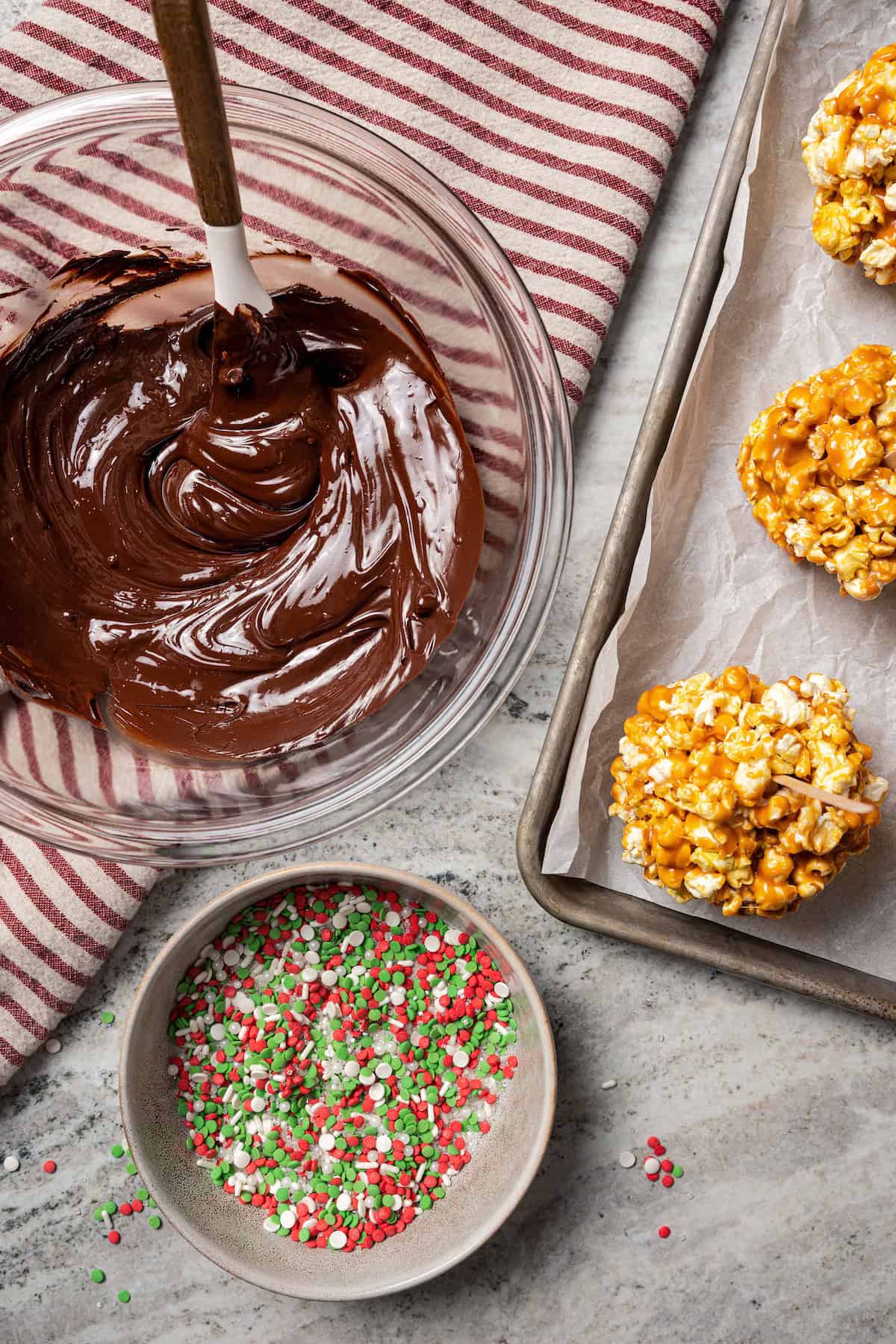 Overhead view of a bowl of melted chocolate next to caramel popcorn balls on a baking sheet and a small bowl of sprinkles.