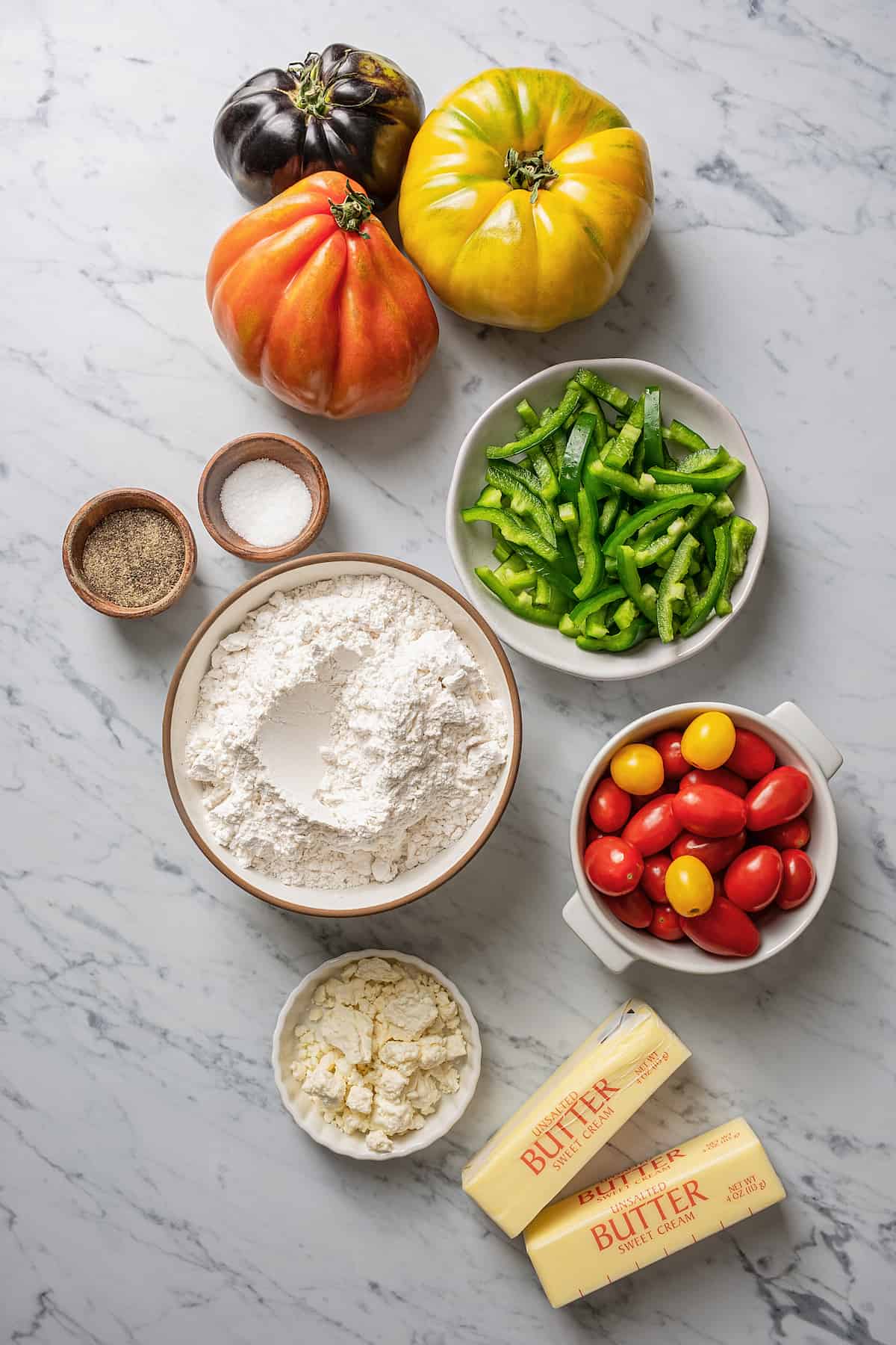 Overhead view of the ingredients needed for a tomato tart: a bowl of flour, a bowl of feta cheese, a bowl of green peppers, a bowl of cherry tomatoes, a bowl of pepper, a bowl of salt, three heirloom tomatoes, and two sticks of butter