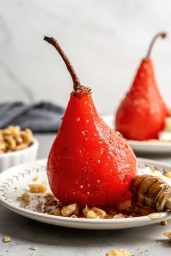 A poached pear on a plate with walnuts and a wooden honey spoon, with a bowl of walnuts and another poached pear in the background