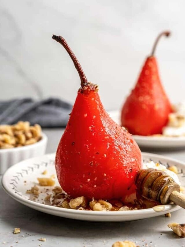 A poached pear on a plate with walnuts and a wooden honey spoon, with a bowl of walnuts and another poached pear in the background