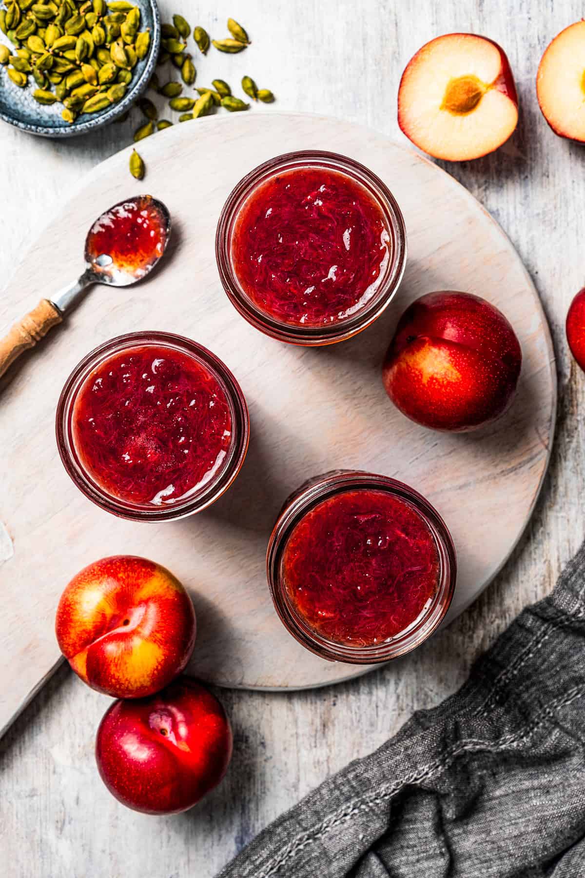 Three jars of jam set on a table with plums scattered around them.