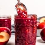 A hand lifting a spoon from a jar of plum jam with fresh plums in the background.