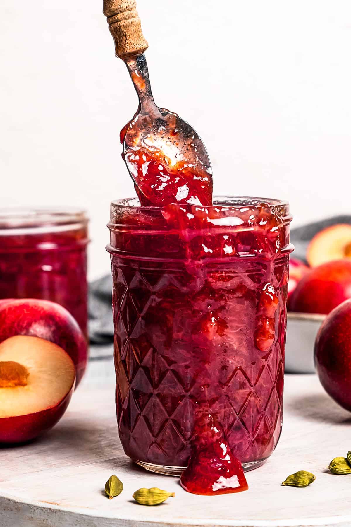 A hand lifting a spoon from a jar of plum jam with fresh plums in the background.