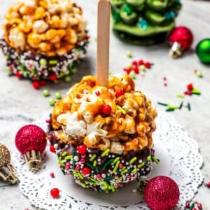 Caramel popcorn balls on a popsicle stick on a paper doily, with a second popcorn ball and a Christmas tree decoration in the background.