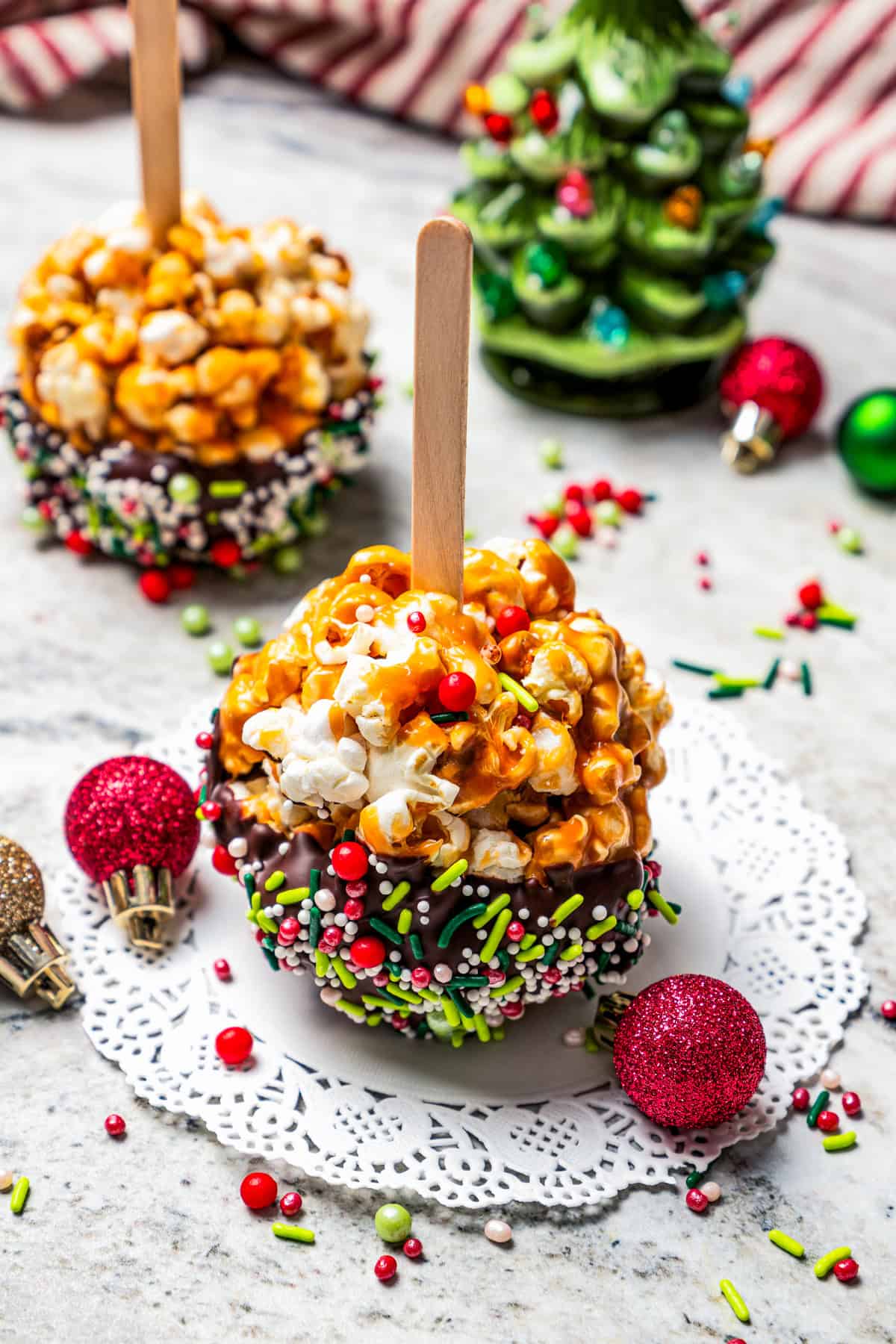 Caramel popcorn balls on a popsicle stick on a paper doily, with a second popcorn ball and a Christmas tree decoration in the background.