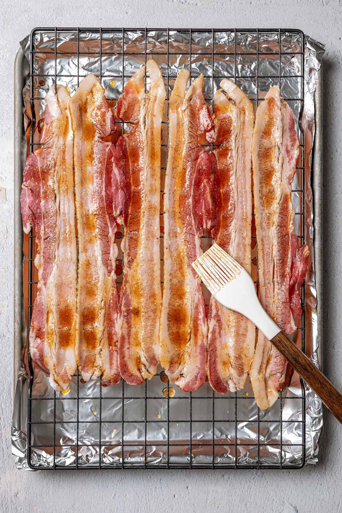 A rubber basting brush brushing sugar and seasonings over bacon strips on a wire rack set over a baking sheet.