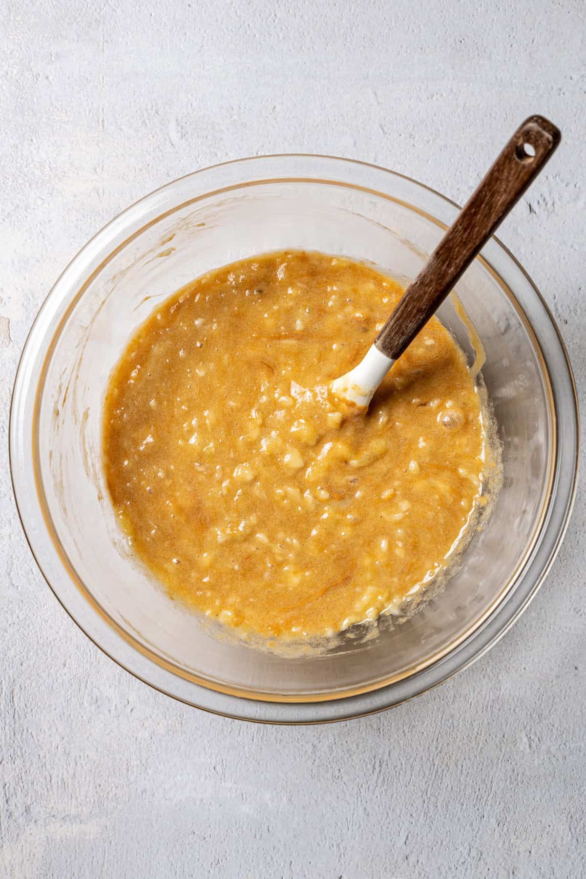 Wet banana bread ingredients combined in a glass bowl with a spoon.