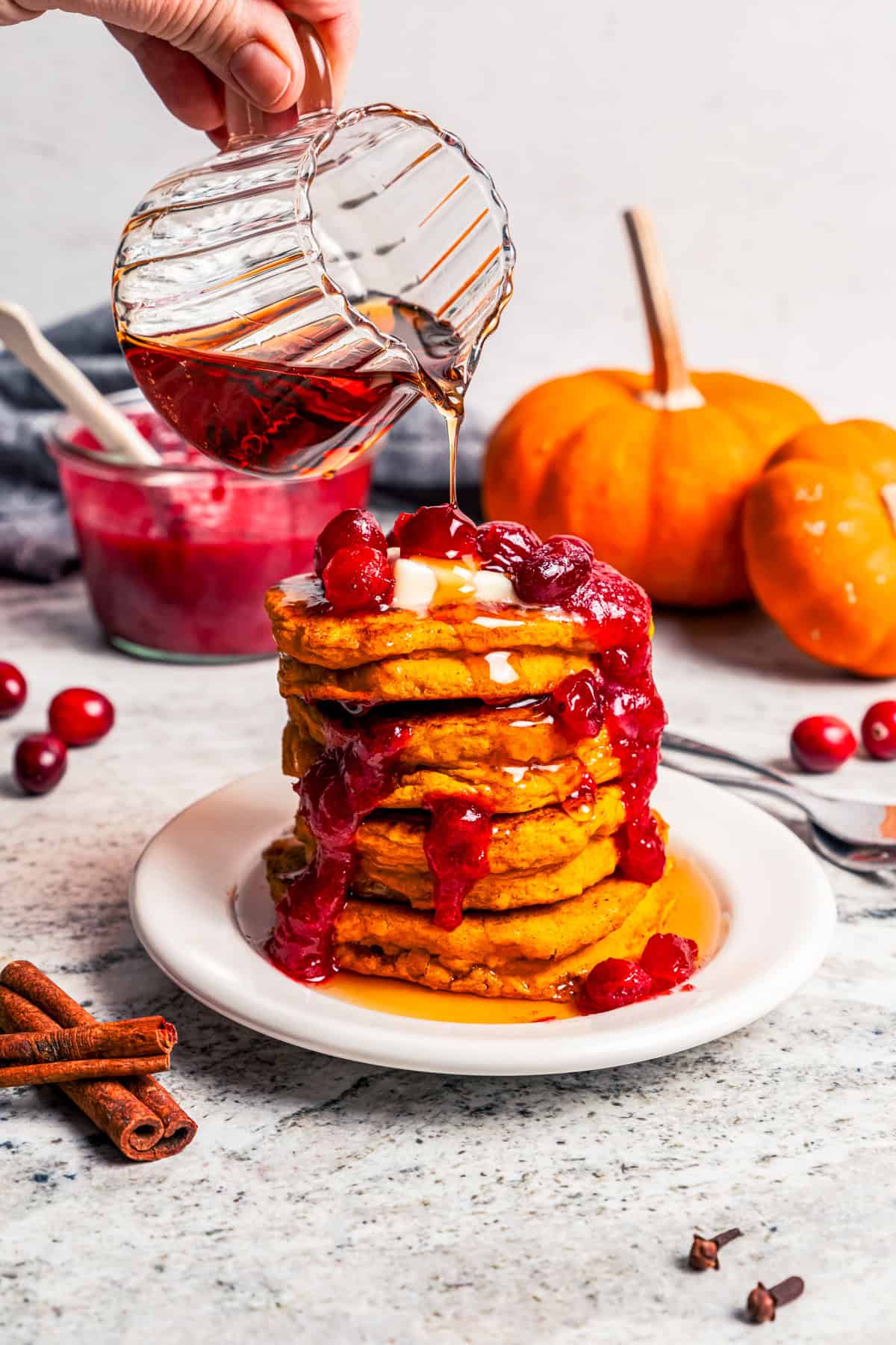 Pumpkin pancakes are the best fall breakfast! Made from pancake mix, real pumpkin, and spices, topped with cranberry syrup for the holidays. Pouring cranberry maple syrup over a tall stack of pumpkin pancakes on a plate.