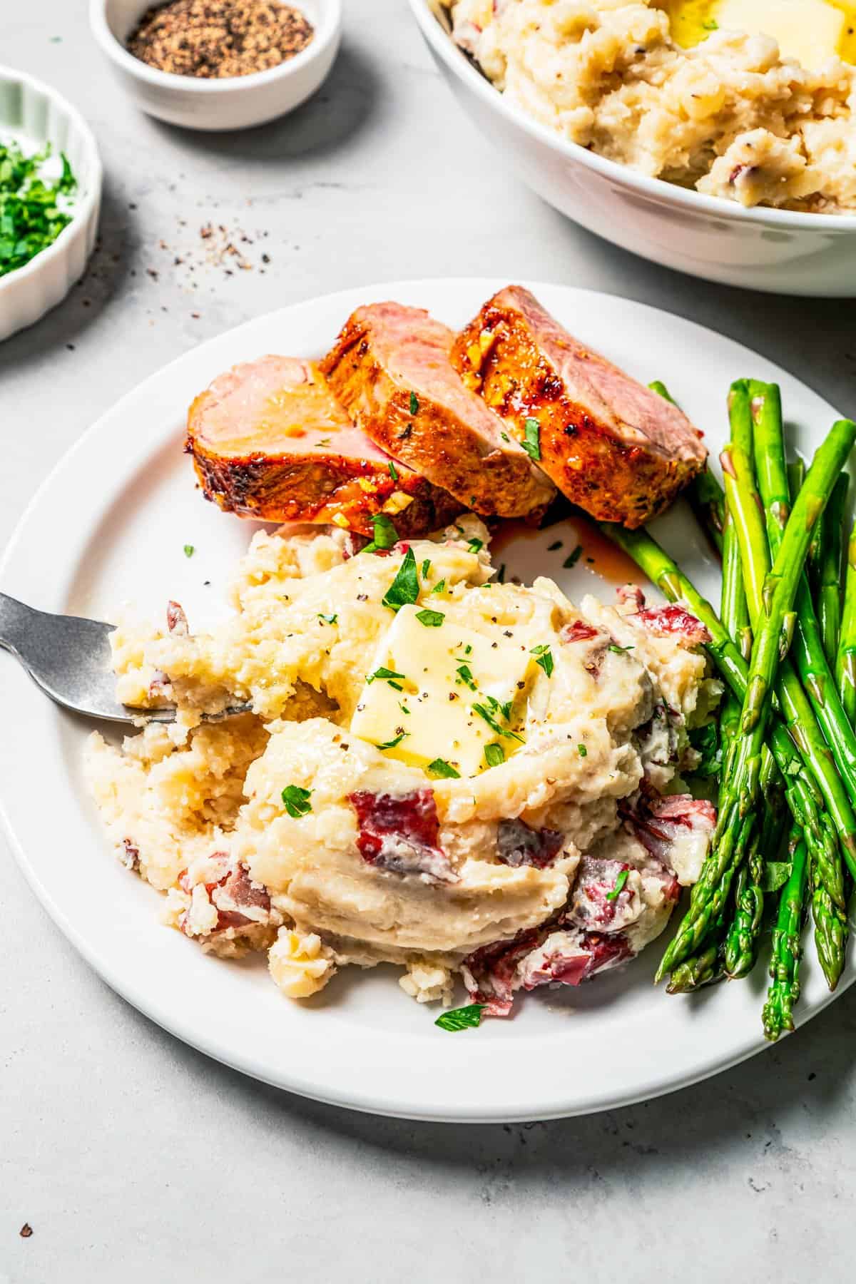 These slow cooker garlic mashed potatoes are made with creamy, skin-on red potatoes and plenty of butter! No boiling, and only 6 ingredients. Country garlic mashed potatoes served next to pork slices and asparagus on a plate.