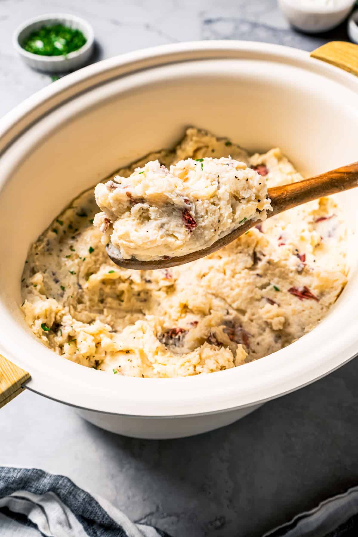 These slow cooker garlic mashed potatoes are made with creamy, skin-on red potatoes and plenty of butter! No boiling, and only 6 ingredients. A wooden spoon scooping country garlic mashed potatoes from the slow cooker.