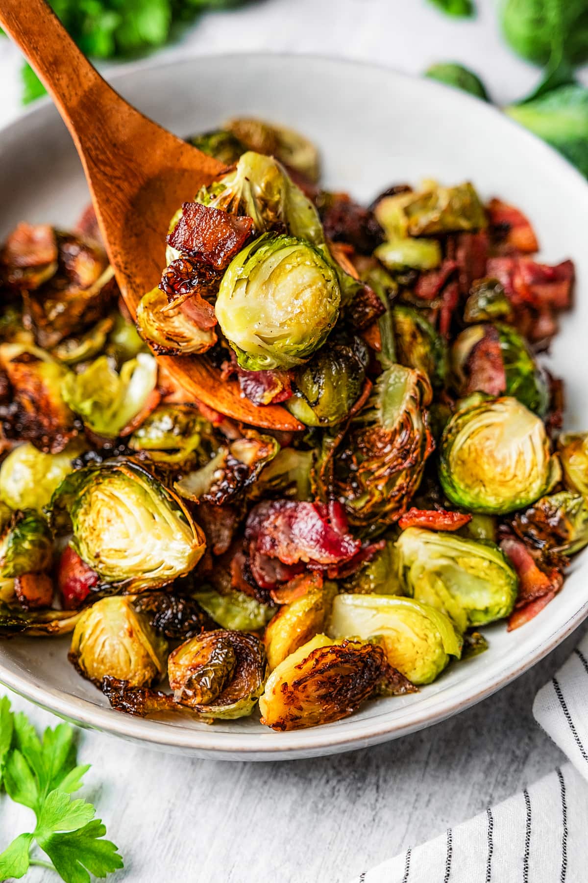 Scooping roasted Brussels sprouts and bacon from a serving bowl with a wooden serving fork.