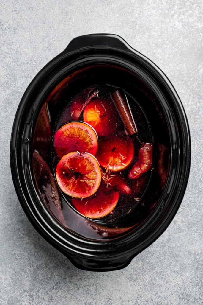 Mulled wine inside the slow cooker with slices of oranges and cinnamon sticks.