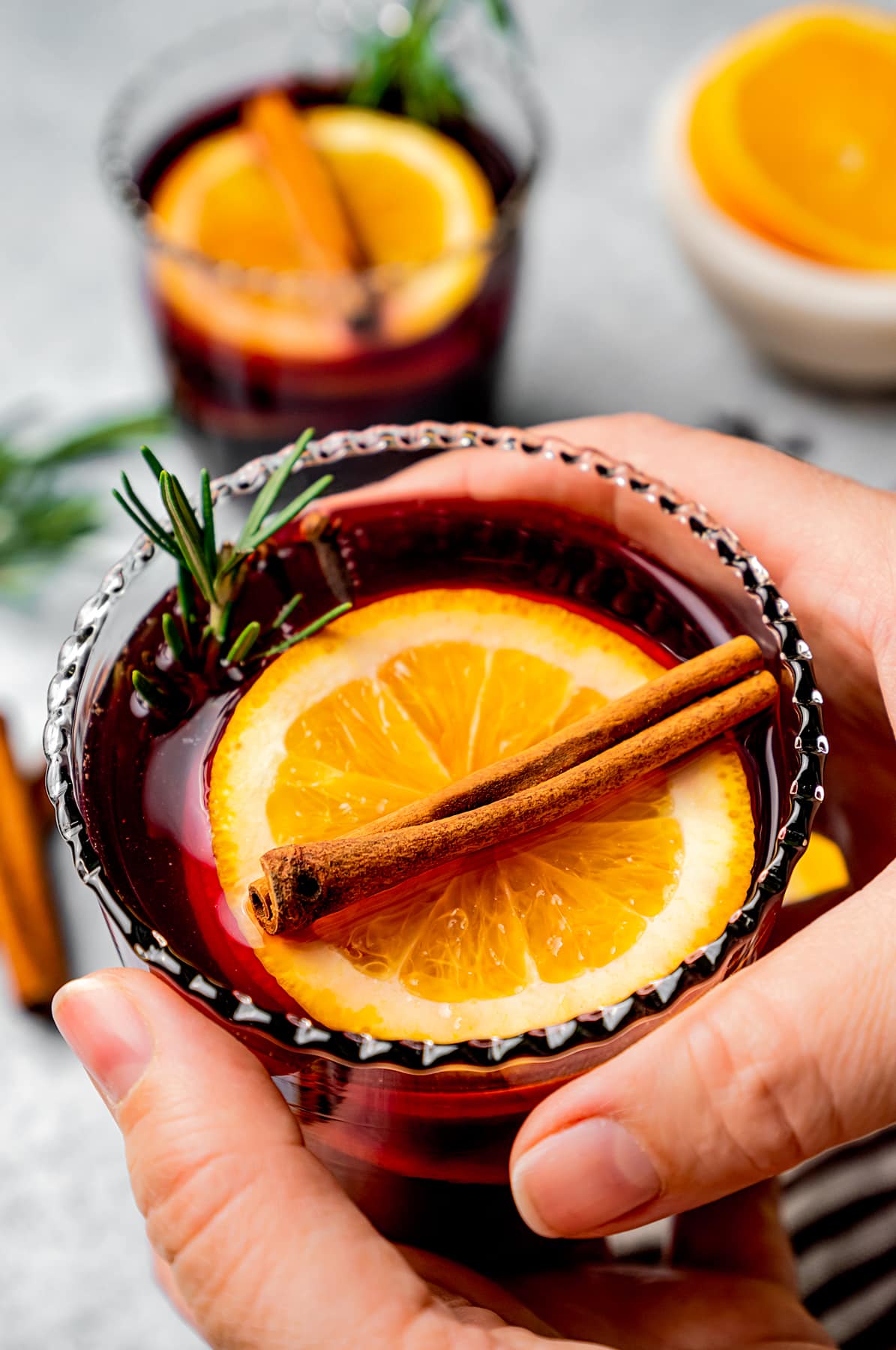 Hands wrapped around a glass of mulled wine garnished with an orange slice and a cinnamon stick.