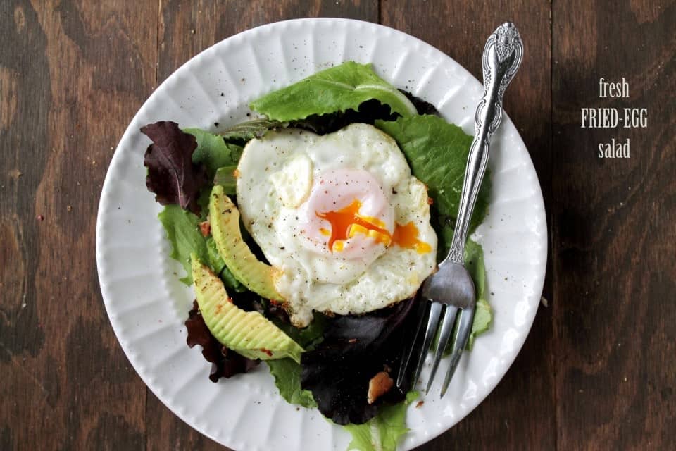 A fried egg with a broken yolk sits on top of a fresh salad with avocado on a white plate.