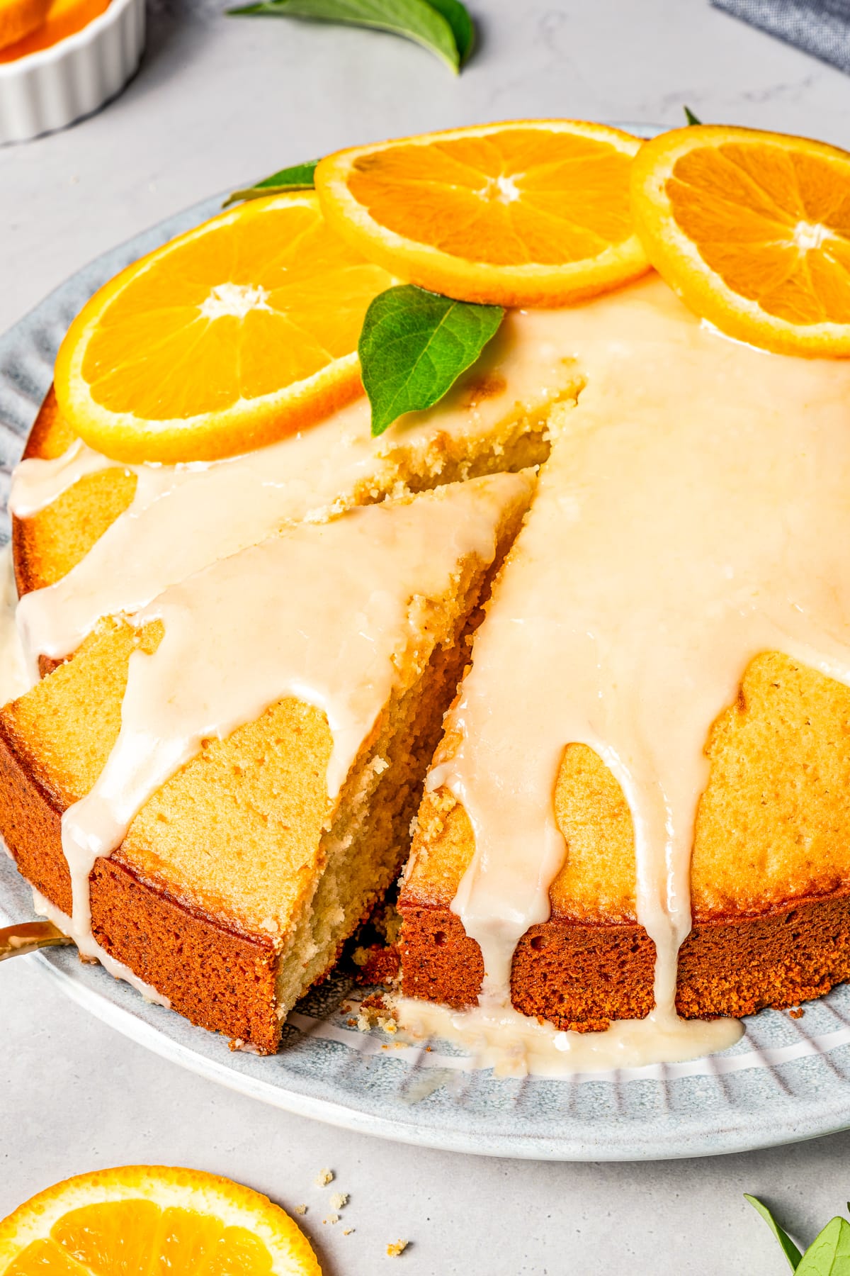 A slice is lifted from a glazed cake garnished with orange slices and mint leaves.